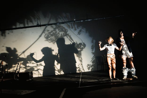 Two performers stand, looking surprised in a dark stage with a projection of flowers illuminating them and the back wall. The performers wear shirts but no pants and have smudges on their faces. 