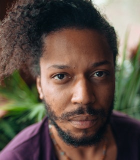 A portrait of Makini (jumatatu m. poe) in a purple shirt and short necklace. They fashion a trimmed beard and their locks in a messy bun on top their head.