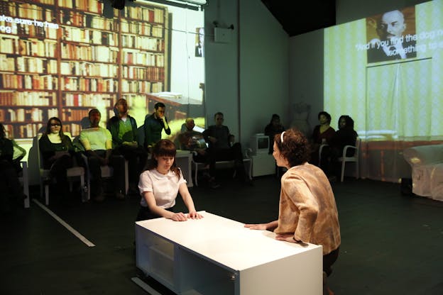 Two performers kneel in front of a low-lying white table surrounded by audience members. One rests their hands on the table, their eyes looking downward as if embarrassed or ashamed. The other has their back to the camera, their hands on the table, looking at the first performer. An image of shelves filled with books is projected against the wall behind them.