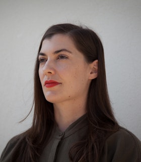 Portrait of Milka Djordjevich with long brown hair and a red lipstick, dressed in a dark green dress shirt looking towards the distance.