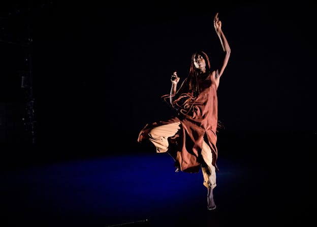 A performer looks upwards, mid-jump with one leg extended and the other lifted and bent. One arm is raised above their head and the other is bent by their side. They wear clothing in various shades of rust-red and orange. A puddle of blue light is beneath them but the rest of the setting is black.