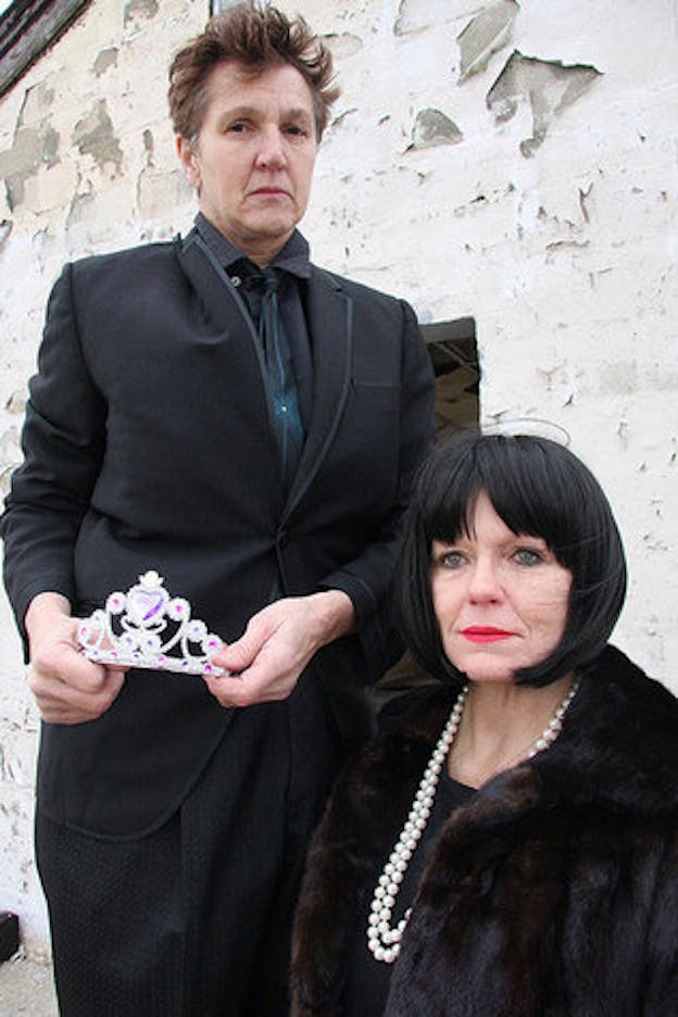 Two performers stand in front of a wall covered in peeling white paint. Peggy Shaw stands on the left. She has short brown hair and wears an all black suit. She holds a plastic silver and pink tiara at her waist. On the right of the image there is an older person sitting down. They have a black bob hairstyle and wear a black top, a pearl necklace, a thick fur coat and red lipstick.