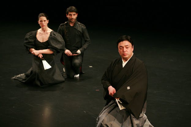 A performance still of three performers sitting on their knees on a black floor. They all look forward, somewhat forlornly and hold fans in their laps. The performer in the foreground wears a kimono. One performer in the background wears a black dress with large puffed sleeves, the other performer in the background wears a jacket with epaulettes and matching pants.  