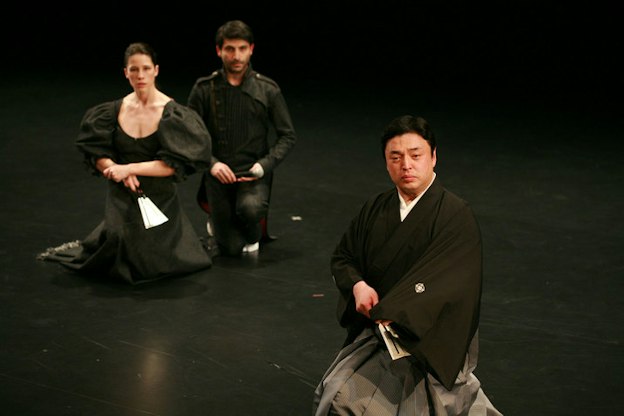 A performance still of three performers sitting on their knees on a black floor. They all look forward, somewhat forlornly and hold fans in their laps. The performer in the foreground wears a kimono. One performer in the background wears a black dress with large puffed sleeves, the other performer in the background wears a jacket with epaulettes and matching pants.  