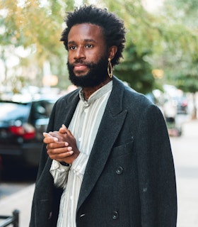 A portrait of JJJJJerome Ellis wearing a gray wool coat, a light blue and white striped shirt, and a large gold hoop earring. He stands in front of a tree-lined street. His body is angled as he stares beyond the camera. He clasps his hands together. 