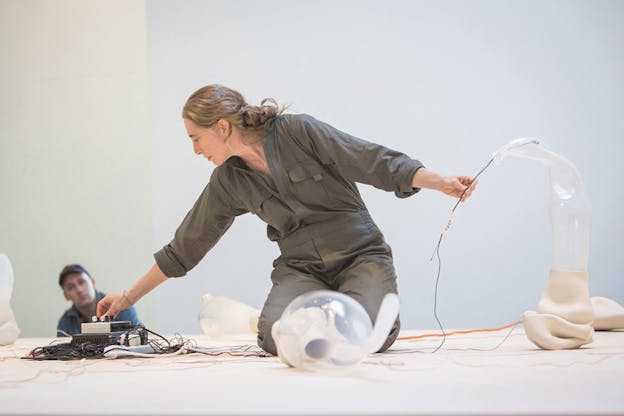 Photograph a person dressed in overalls kneeling on the floor surrounding by plastic white and transparent materials connected together. In one hand she touches the controls of a small electric box machinery while on the other she holds the cables of that machinery in one of the artist materials.