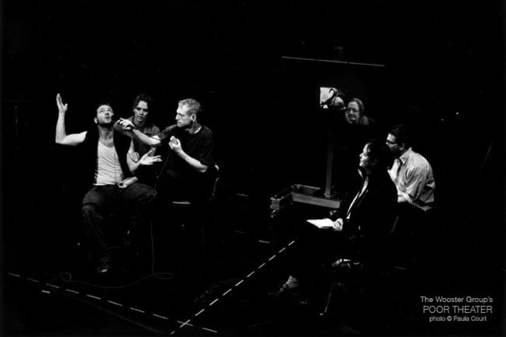 Three people sit on the left side within a space demarcated on the ground by a surrounding dotted line. One person holds their arm up and looks up while the two other people look intently at them and one person holds out a microphone for them. On the right, three people watch from chairs. 