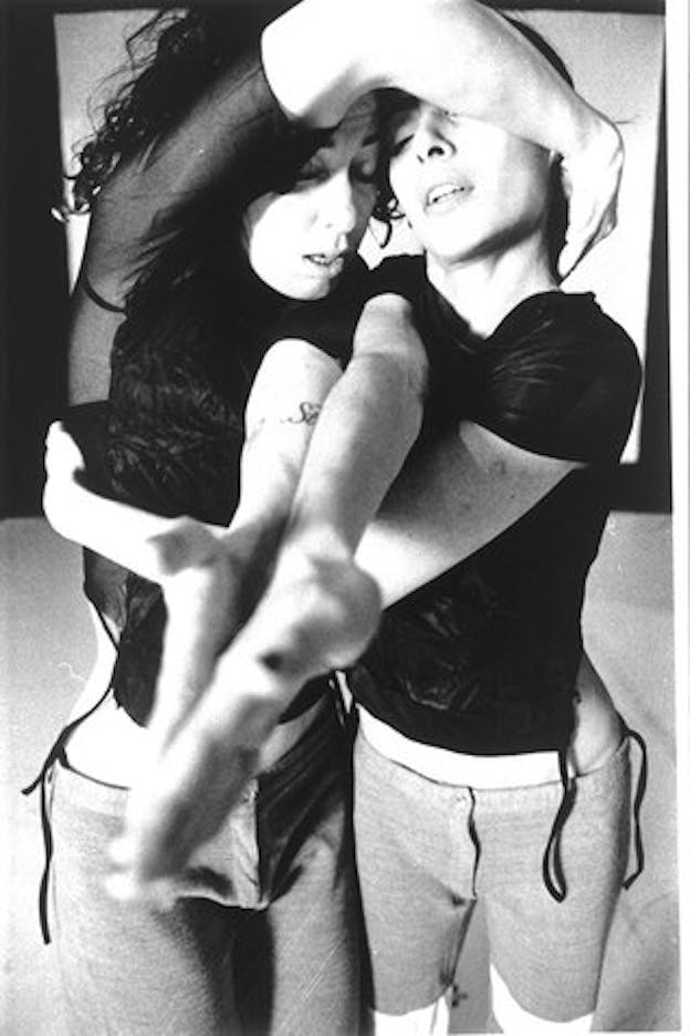 A close up black and white image of two performers intertwined. They both wear black mesh tops and short cotton pants. Both of them extend one arm forward to touch, using their other arm to wrap around the other performer.  