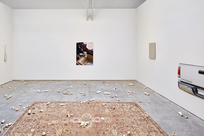 Image of a kitchen sink with cups, greens and a mask to its side, hangs on a wall surrounded at its bottom by bone fragments on a carpet.   To the right a cut out of a cars back hangs on a wall. 