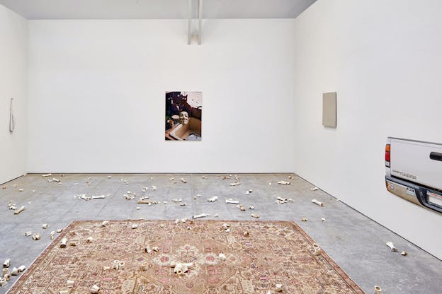 Image of a kitchen sink with cups, greens and a mask to its side, hangs on a wall surrounded at its bottom by bone fragments on a carpet.   To the right a cut out of a cars back hangs on a wall. 