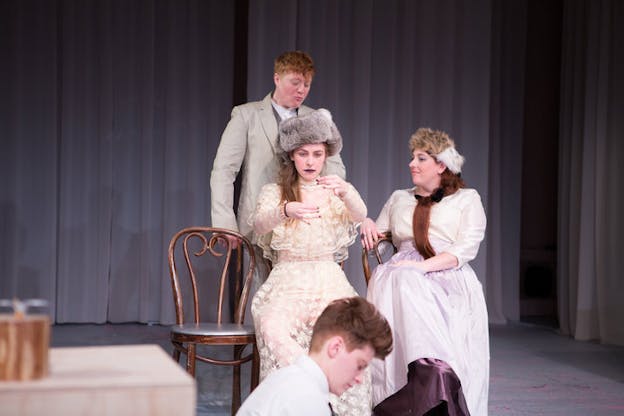 A performer dressed in a lace dress with a gray fur hat sit on a chair with their hands raised in the front. Next to them a figure dressed in a cream shirt and lilac skirt with a beige fur hat touches their shoulder. Above them a third person dressed in a gray suit stares. 