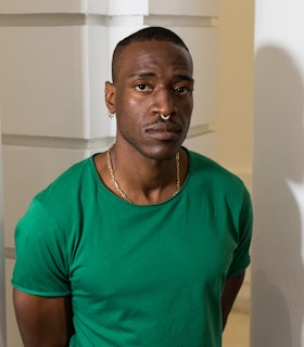 A portrait of Carlos Martiel wearing a green t shirt, a golden necklace chain, small gold hoop earrings, and a gold nose ring. He has short hair and stands in front of a white background. 