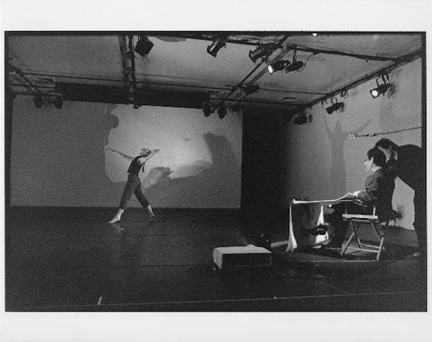 A performer in front of a shadowy screen steps forwards and bends their back backwards. In the right foreground, two other people watch from directors folding chairs. 