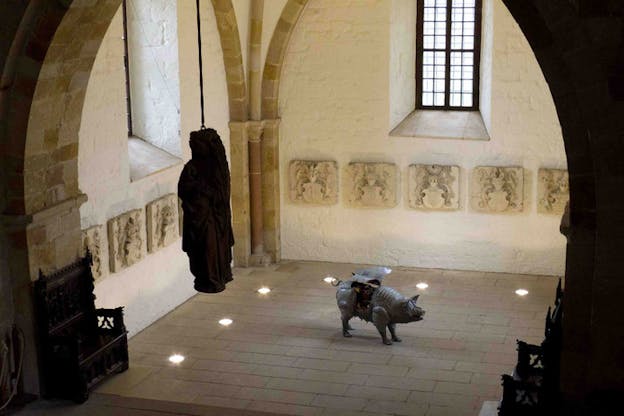 An installation image of an ornate pewter pig on the stone floor of a chapel. Two doors on the pigs mid-section are opened, revealing some machinery inside. Above the pig, a stone sculpture of a cloaked figure hangs by a rope from the ceiling. On the chapel walls, numerous ornate tiles decorated with coats of armor are aranged in a horizontal line. All of this is visible through a archway and while the room with the pig is brightly lit by two windows, everything beyond the arch is shrouded in shadows.