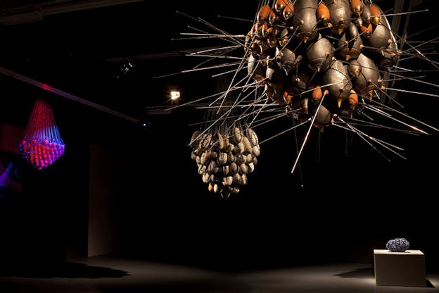 Three round structures consisting of smaller oval shapes hang from a ceiling. The structure in the front closest to the viewer contains silver and orange metallic ovals with antennae stuck on them. The structure behind the first holds marbled white ovals. The third structure farthest from the viewer to the left holds red ovals illuminated by blue colors.