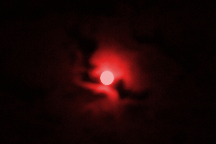 Blurred image of a white sun surrounded by neon red clouds fading into black sky.