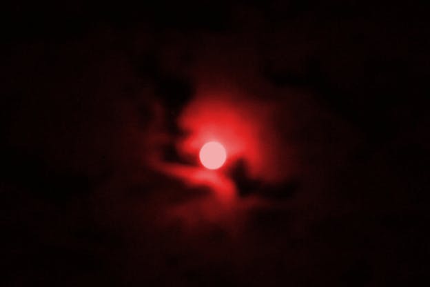Blurred image of a white sun surrounded by neon red clouds fading into black sky.