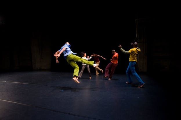 Performers dressed in bright slacks and shirts leap and twist on an empty dark stage. 