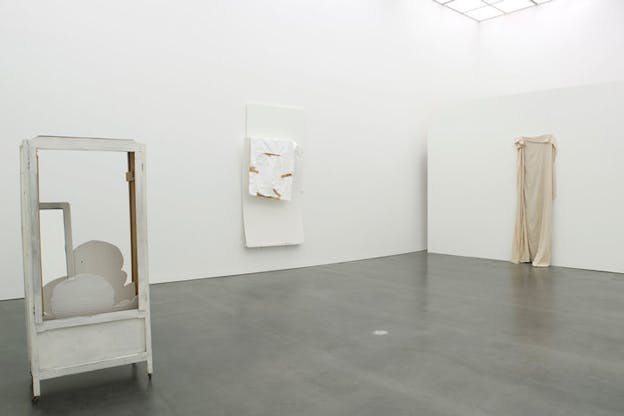 An installation image of an exhibition room with white walls and grey floors. On the left, there is a vertical, rectangular frame, painted white. On the bottom of the frame, there are thin curved white objects that look like clouds. On the back wall, there are two overlapping large pieces of paper with a thin sheet of tissue paper covered with bits of brown tape resting atop. On the right wall, a thin, long, beige sheet hangs from the wall. 