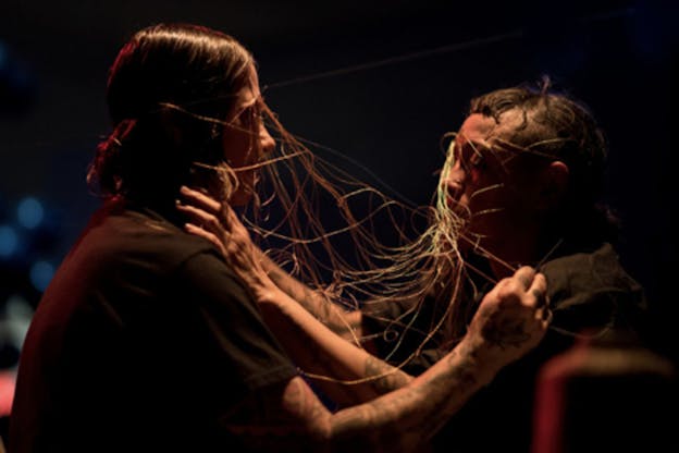 Two people facing each other in dim lighting with thin cables on their faces that meet and tangle in the space between them, each extend their arms towards each other holding on the cables.