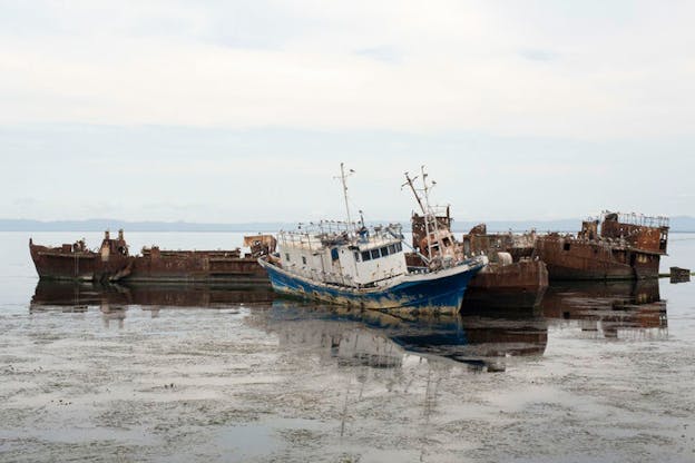 A rusting blue and white fishing boat floats against two competely rusted barges in a still, overcast bay. 