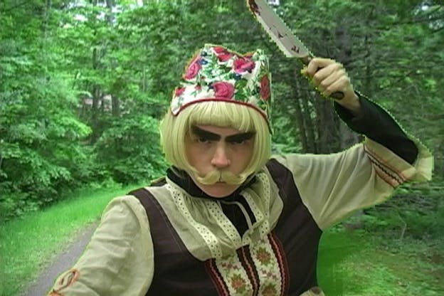 A video still of Ben Tor wearing a short blonde wig, bushy eyebrows, a blonde handlebar mustache, a rose pattern hat, and an ornate blouse. She stands in a forest and weilds a bloody knife above her head. Her arm and the knife are outlined by a thin pixelated yellow line. 