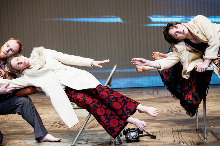 Performers leaning sideways on chairs, dressed in fur-lined coats. One performer wearing ornately patterned scarlet and navy dress pants is being held by a performer kneeling on the wooden floor.
