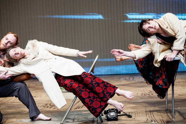 Performers leaning sideways on chairs, dressed in fur-lined coats. One performer wearing ornately patterned scarlet and navy dress pants is being held by a performer kneeling on the wooden floor.