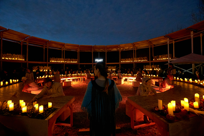 A person dressed in black at the center of the image with their back turned to the viewer watches people dressed in beige as they sit around tables that form a circle. They are situated in an open dome theater,lighted by multiple candles in every corner. Across the person dressed in black the word 'feast' shines in white neon light.