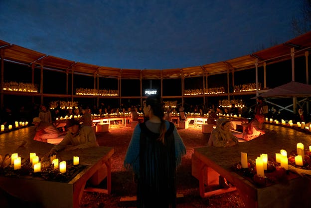 A person dressed in black at the center of the image with their back turned to the viewer watches people dressed in beige as they sit around tables that form a circle. They are situated in an open dome theater,lighted by multiple candles in every corner. Across the person dressed in black the word 'feast' shines in white neon light.