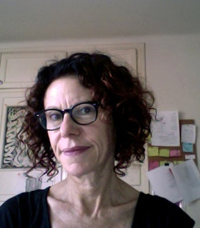 Portrait of Amy Gerstler short curly dark auburn hair, dressed in a black shirt and black thick framed glasses. 