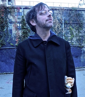 Image of Anselm Berrigan in jeans and a black coat looking up sideways while holding a tiger plushie in their pocket.
