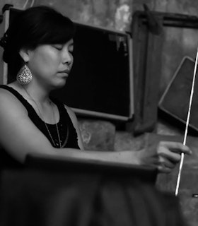 A black and white profile of Bonnie Jones holding a metal rod, looking downward. The artist is wearing a black sleeveless top, a large teardrop-shaped earring, and has their hair swept across the side of their face.