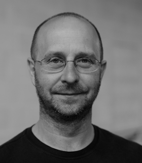 A black and white portrait of Jonathan Burrows wearing a dark top and wire rimmed glasses. He looks directly at the camera and smiles slightly. The background is blurred.