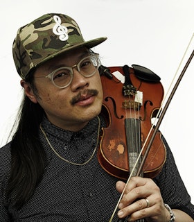 Portrait of Spencer Yeh holding a violin and bow over the shoulder, wearing a camouflage print hat, clear glasses, and a black button down shirt.