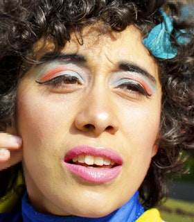 Close up portrait of Larissa Velez-Jackson with curly dark hair, wearing vibrant pink and blue eye makeup.