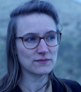 Close up portrait of Catherine Lamb with their hair pulled back, wearing round brown glasses and staring into the distance.
