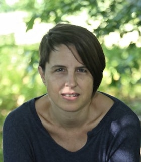 Portrait of Mallory Catlett hunched over toward the camera with side swept short hair and a dark top. Behind them is a blurred background of a tree.