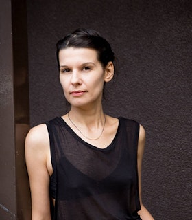 Portrait of Clarissa Tossin leaning with one shoulder against the wall. The artist is dressed in a black sheer top, while their straight black hair are cleared from the front.