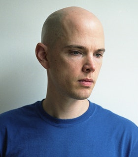 Portrait of Chris Corsano in a blue shirt with blue eyes and bald looking sideways.