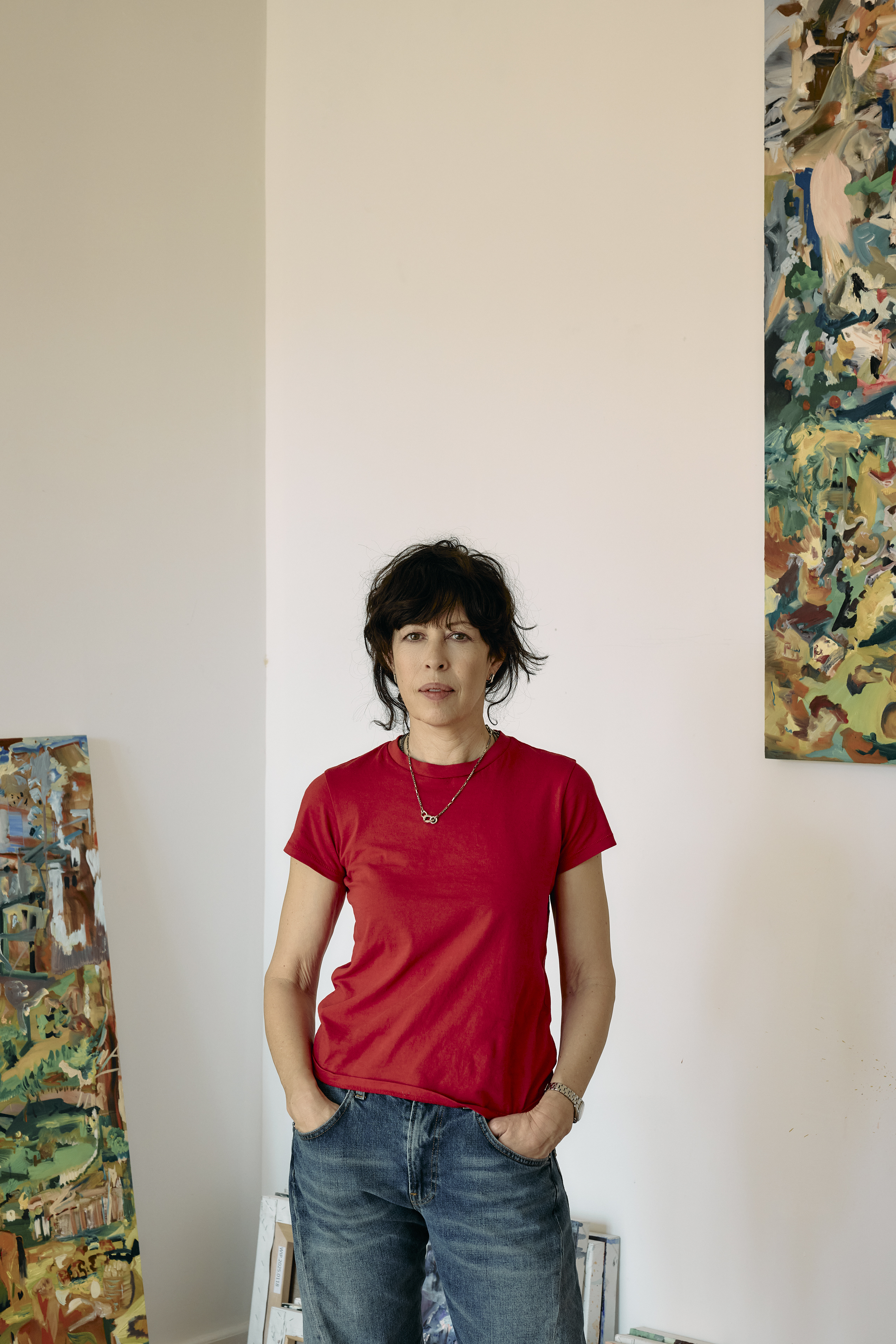 Cecily Brown in her studio standing with her hands in her pockets and wearing a read t-shirt and jeans. Partial views of two of her paintings are visible behind her, to the left resting on the floor, and to the right hanging on a wall.