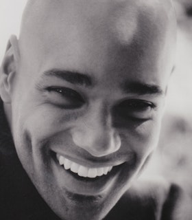 Black and white portrait of Darrell Jones wearing a turtleneck, bald with thickly shaped brows smiling.
