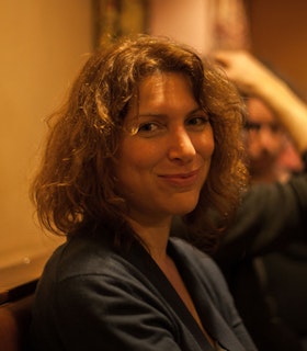 Annie Dorsen smiling at the camera with medium length wavy hair in yellow lighting.