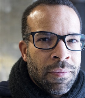 Close up portrait of Douglas Kearney with a black scarf surrounding the neck, black thick framed glasses and a beard.