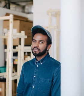 Portrait of EJ Hill wearing a blue hat and a denim button up shirt in front of blurred wooden structures.