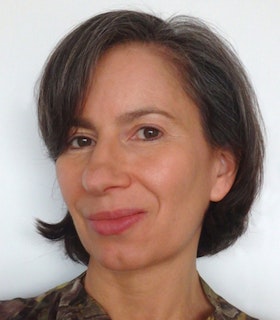 Portrait of Andrea Fraser smiling at the camera with short brown hair in front of a white wall.