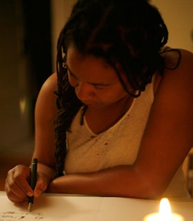 Renee Gladman looks down and writes on a paper in a candlelit space.