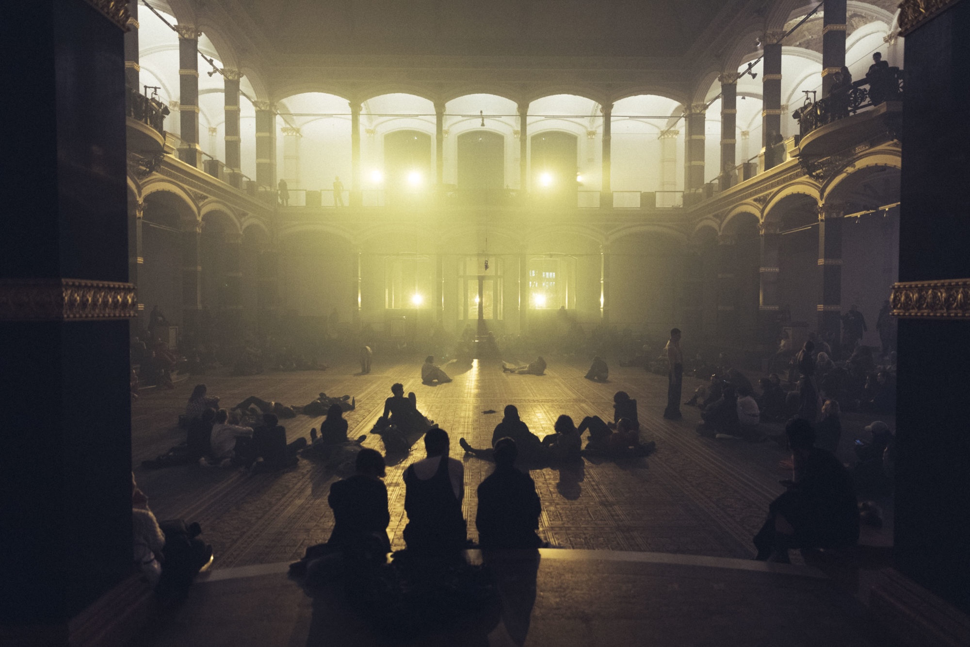 Pan Daijing performing in an open floor plan concert hall space with a series of collaborators. Soft yellow lights shine out from the back of the hall, illuminating the space in soft light. The audience is seen sitting and lying down throughout the space, some on the ground floor and others on the balcony.