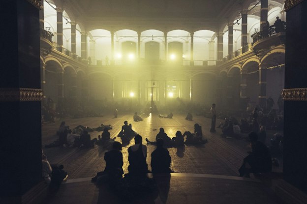 Pan Daijing performing in an open floor plan concert hall space with a series of collaborators. Soft yellow lights shine out from the back of the hall, illuminating the space in soft light. The audience is seen sitting and lying down throughout the space, some on the ground floor and others on the balcony.