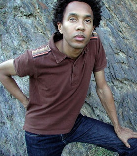 A portrait of Jace Clayton in front of a textured rock wearing a brick red short sleeved polo with stripes on the shoulders and blue jeans. He leans forward, resting on hand on a bent knee and bending the other arm behind his back. 